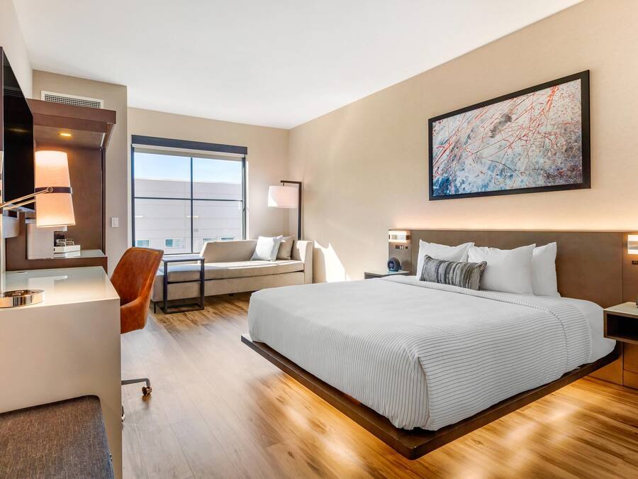 Et moderne hotelværelse med en stor seng, et skrivebord, en stol og en sofa. Der er et stort vindue og et billede på væggen.