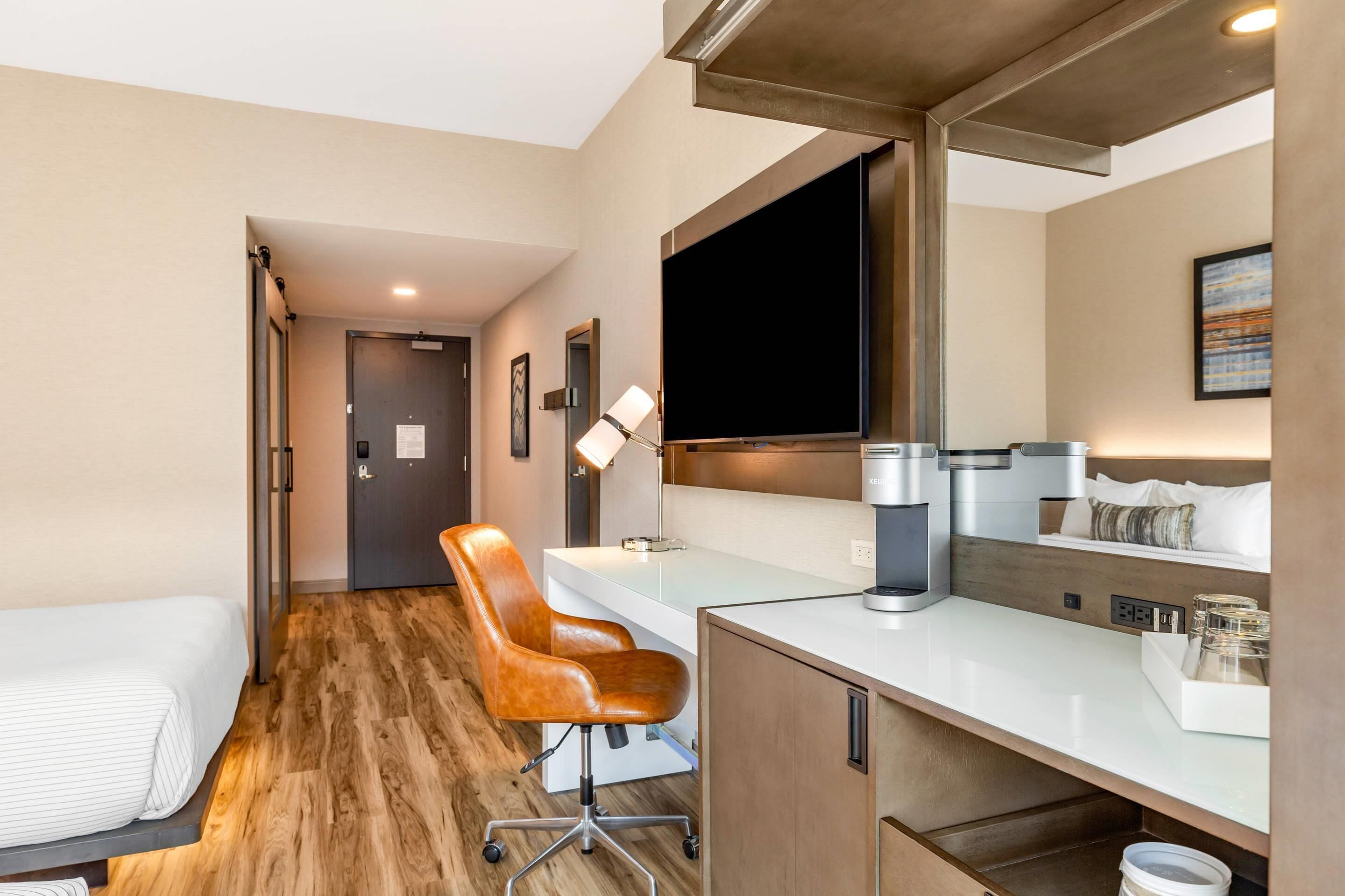 A hotel room with a bed, a desk, a chair, a coffee maker, and a TV.