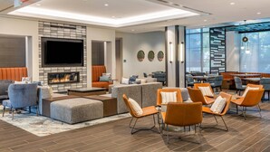 A modern lounge area with a fireplace, seating arrangements, and a large window providing natural light. - Cambria Hotel New Haven University Area (New Haven)
