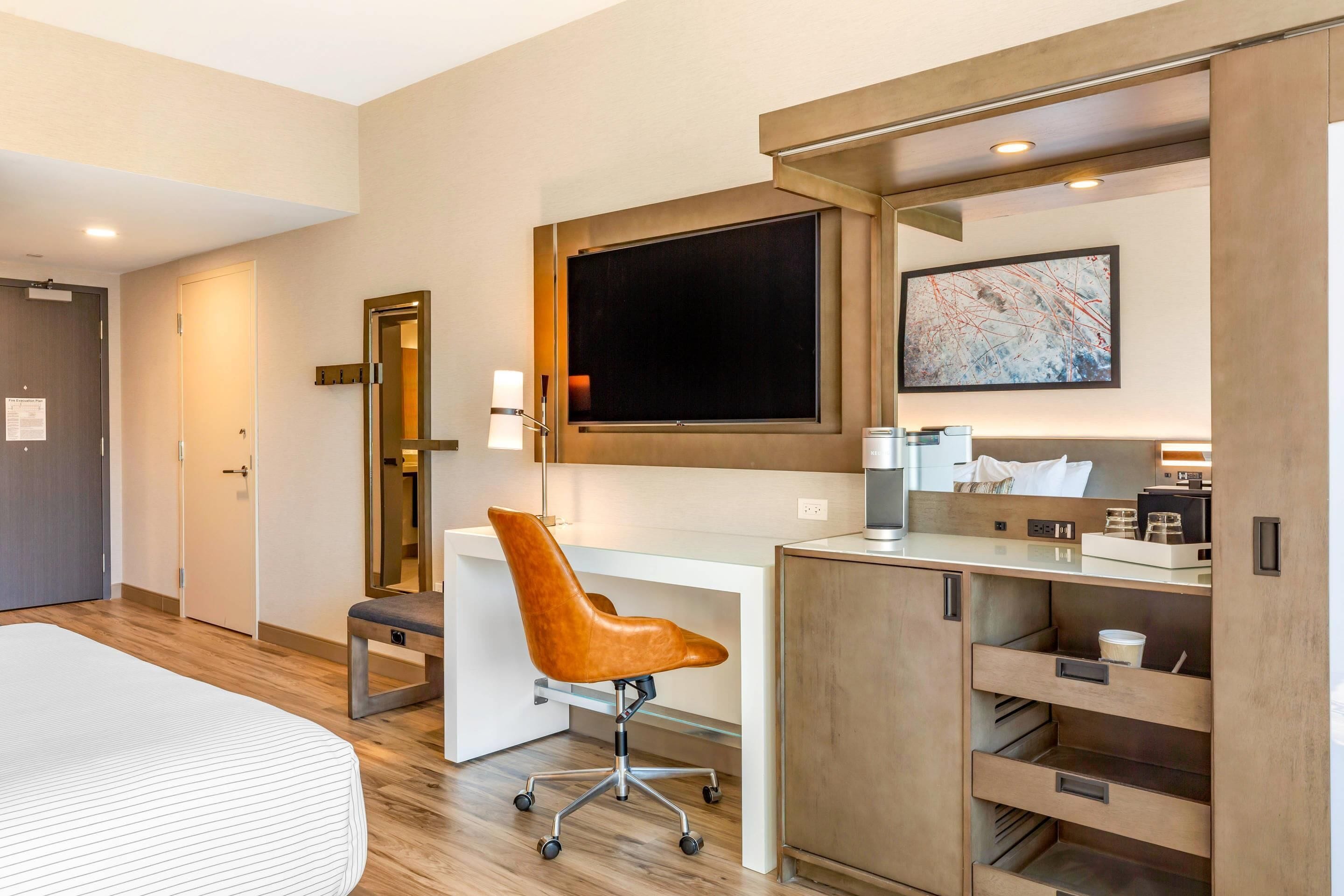 Habitación de hotel con cama, escritorio con silla, televisor de pantalla plana, cafetera y aparador con cajones.