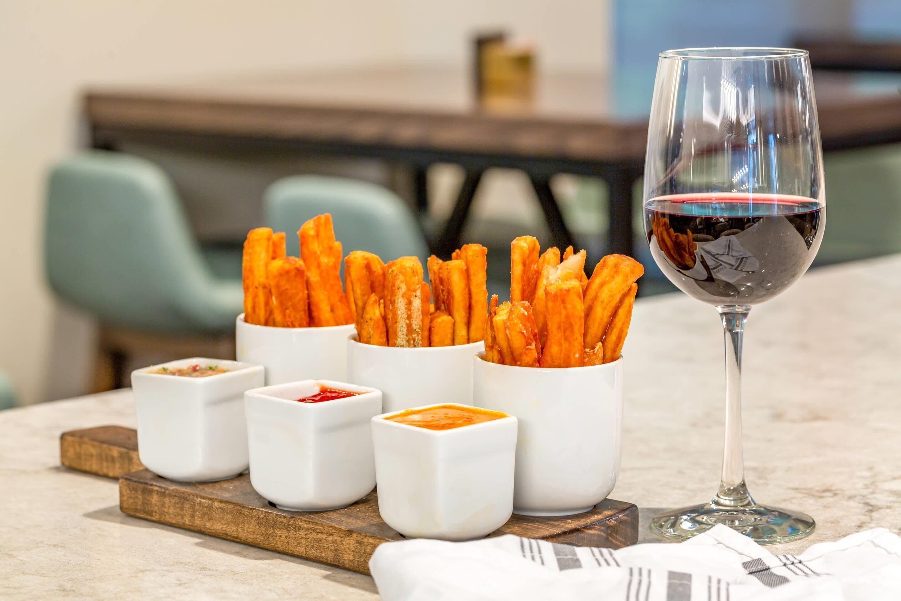 A table with a glass of red wine, a tray with several small white cups containing different sauces, and a container of breadsticks.