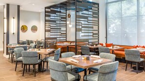 A modern dining area with wooden tables and grey upholstered chairs, a decorative wall with horizontal slats, and large windows offering a view of trees. - Cambria Hotel New Haven University Area (New Haven)
