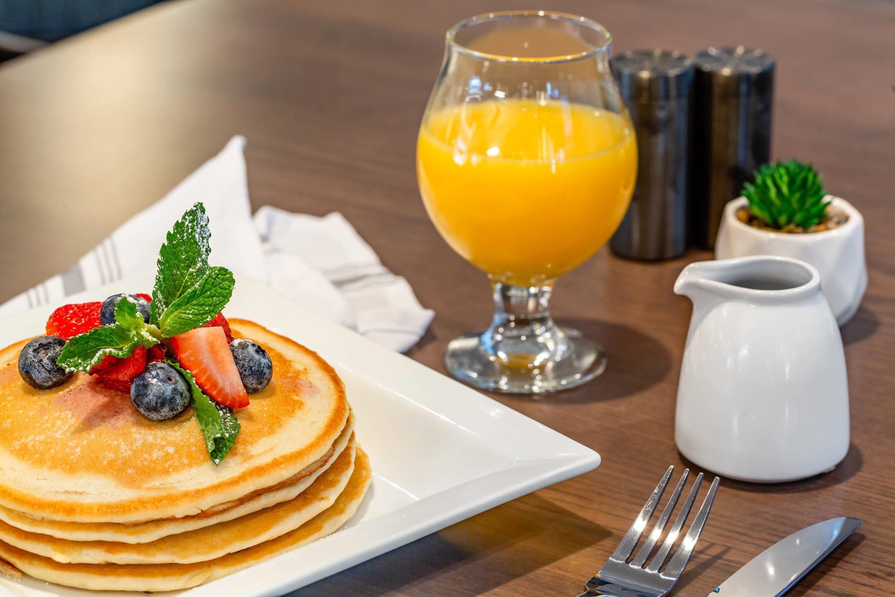 Desayuno con tortitas cubiertas de fresas y arándanos, un vaso de zumo de naranja y una jarra pequeña.