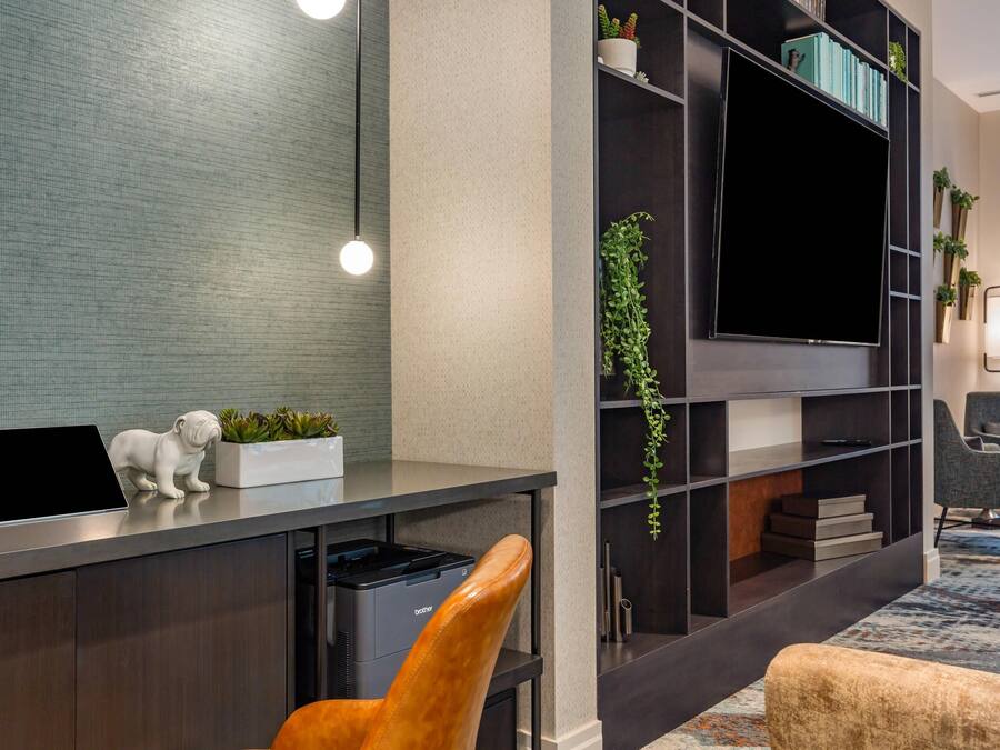 Une chambre d’hôtel moderne équipée d’un téléviseur à écran plat, d’un ordinateur portable, d’une imprimante, d’un canapé, d’un fauteuil et d’une bibliothèque agrémentée de plantes.