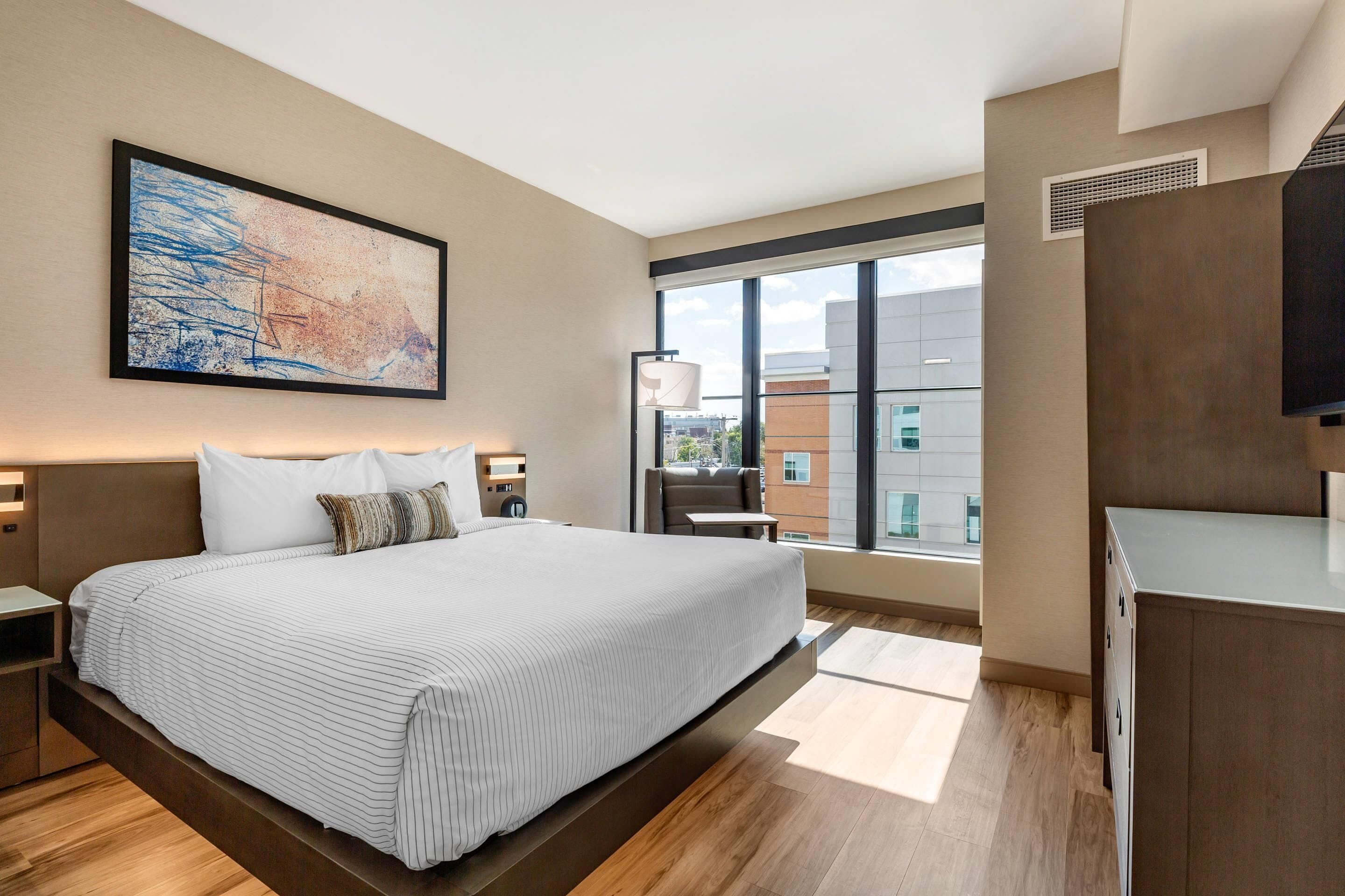 A modern hotel room with a large bed, a painting on the wall, a window with a view of buildings, and a desk with a lamp.