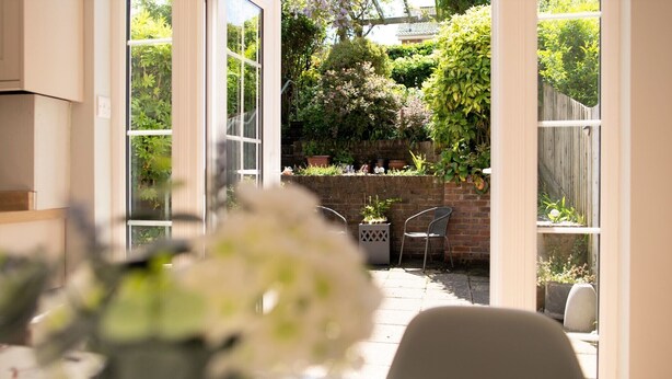 Une terrasse ensoleillée avec une table et des chaises, un mur de briques et une végétation luxuriante en arrière-plan.