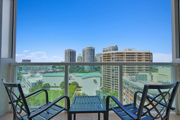 A balcony with two chairs and a table, overlooking a cityscape with high-rise buildings and a green park.