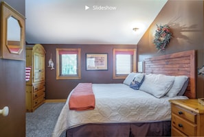 Une chambre à coucher avec une tête de lit en bois, un lit avec une literie blanche et rose, deux fenêtres équipées de stores, une commode et un miroir.