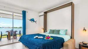 A bedroom with a bed, a dining table with chairs, a view of the sea, and a balcony.