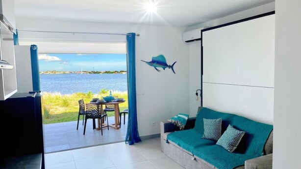 A room with a view of a body of water, a dining area with chairs and a table, and a sofa with patterned cushions.