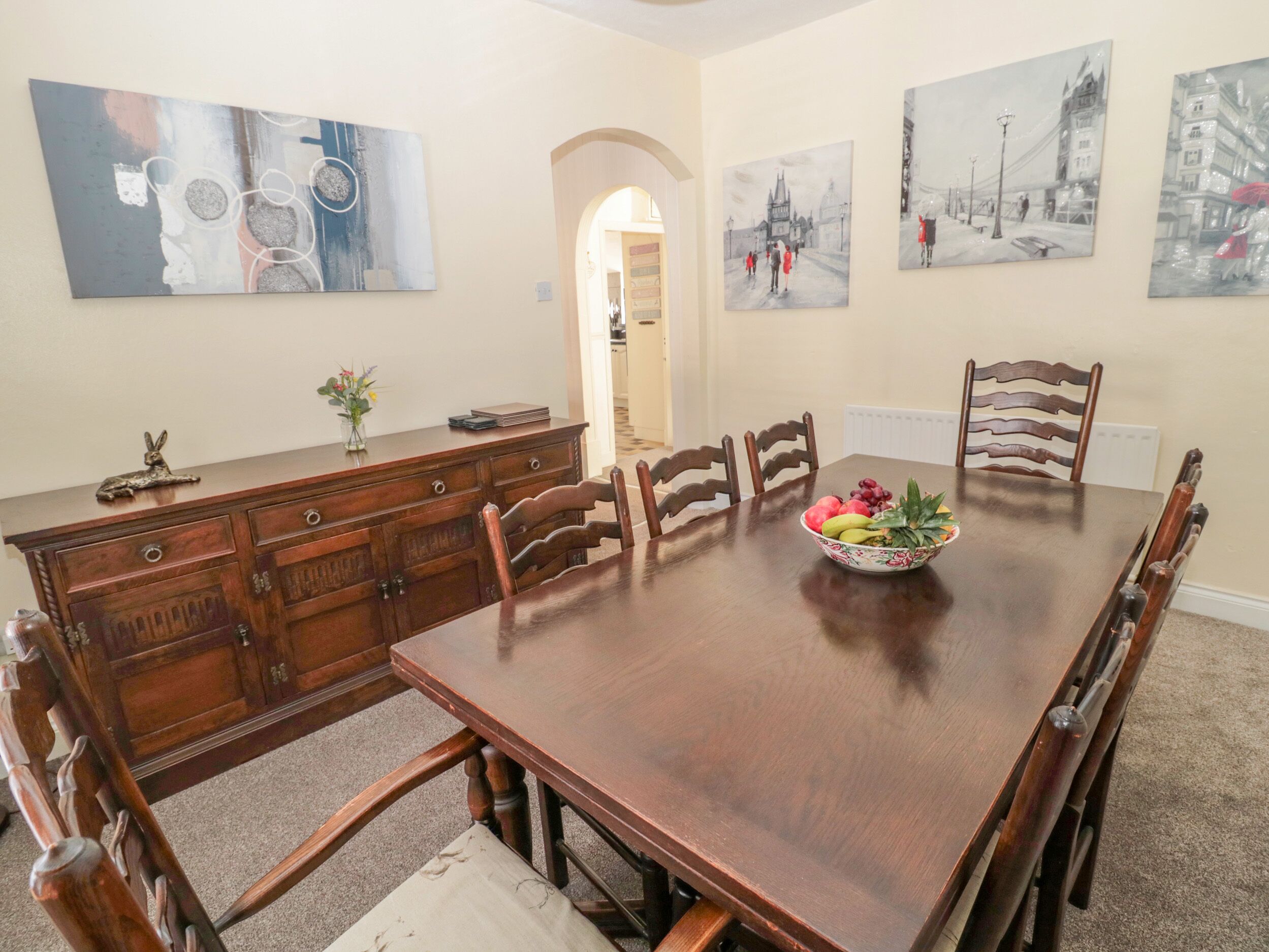 Un comedor con una gran mesa de madera, seis sillas, un florero con flores y tres cuadros en las paredes.