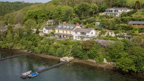 Une grande maison dotée d’un balcon donnant sur une rivière, entourée d’une végétation luxuriante et de collines.