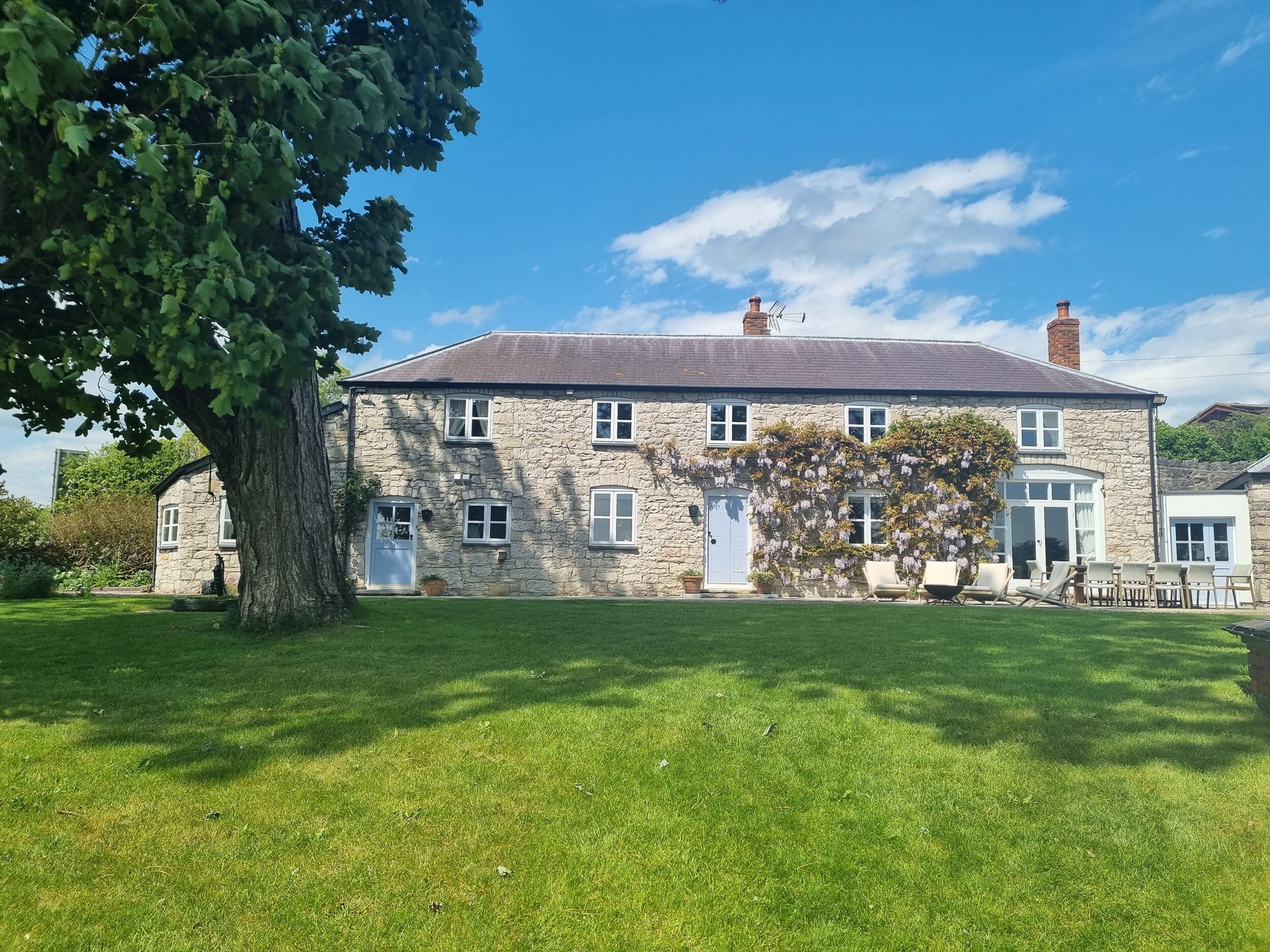A stone house with a large tree in front, a grassy lawn, and a clear blue sky.