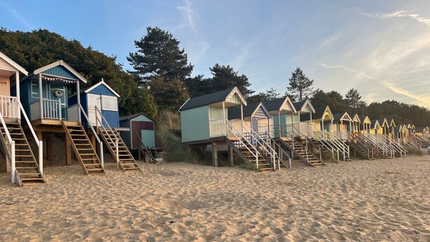 A row of beach huts with wooden stairs leading up to them, set on a sandy beach with trees in the background. - Springfield (Fakenham)