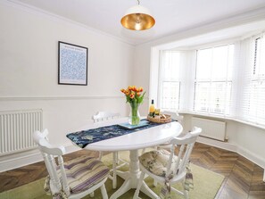 A dining area with a white table, chairs, a vase of flowers, a fruit basket, and a framed picture on the wall.