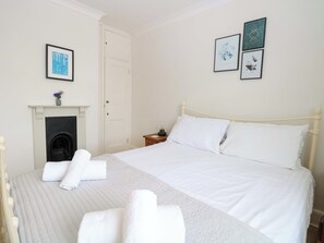 Een keurig opgemaakt slaapkamer met een bed, kussens, een nachtkastje, een bijzettafeltje, een open haard en lijstwerken aan de muur.