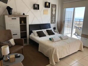 A hotel room with a bed, a desk, a chair, a TV, and a balcony with a view. - Panoramic Holidays - Superior 17 - Modern Apt with PANORAMIC VIEWS  - Sea View (Peyia)