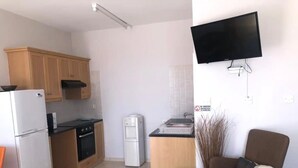 A compact kitchen with wooden cabinets, a refrigerator, a stove, and a microwave. There is a small dining area with a chair and a table. A flat-screen TV is mounted on the wall above the kitchen counter.
