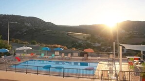 A pool area with lounge chairs and umbrellas, surrounded by a fence, with a mountainous landscape in the background.