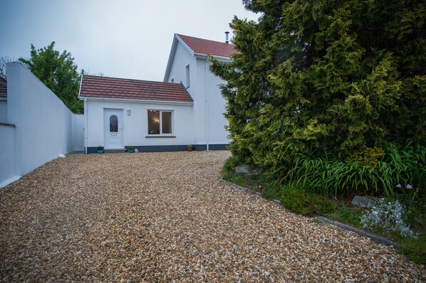 A white building with a red roof, a gravel driveway, and a dense green bush to the right. - Shorelands - 1 Bedroom - Manorbier (Tenby)