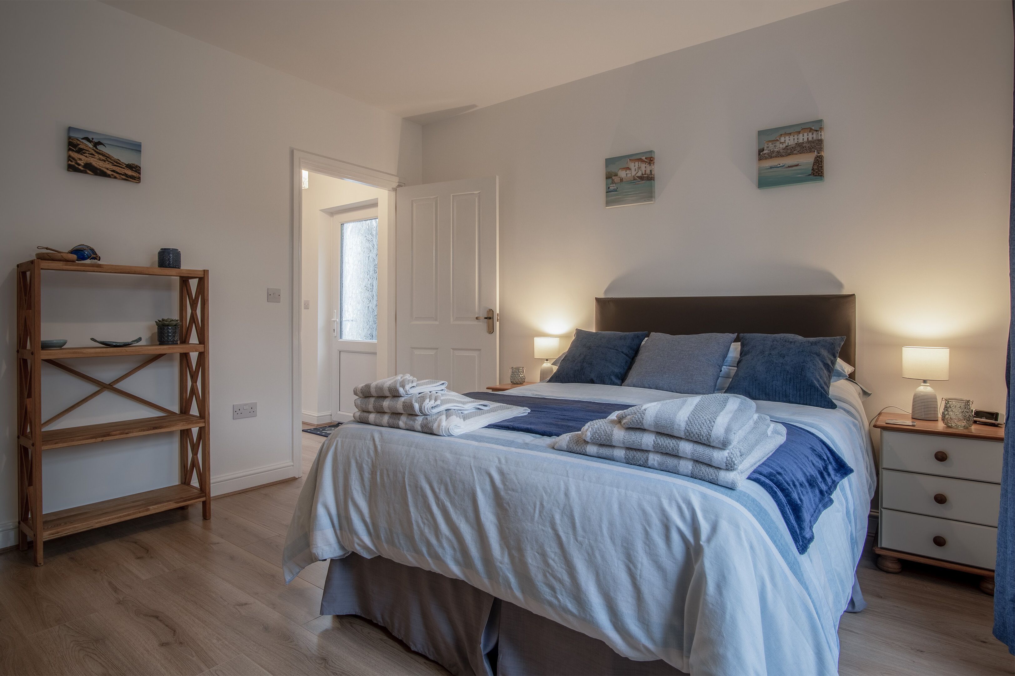 A neatly made bed with a blue and white duvet, a wooden nightstand with a lamp, and a wooden shelf unit against a white wall.