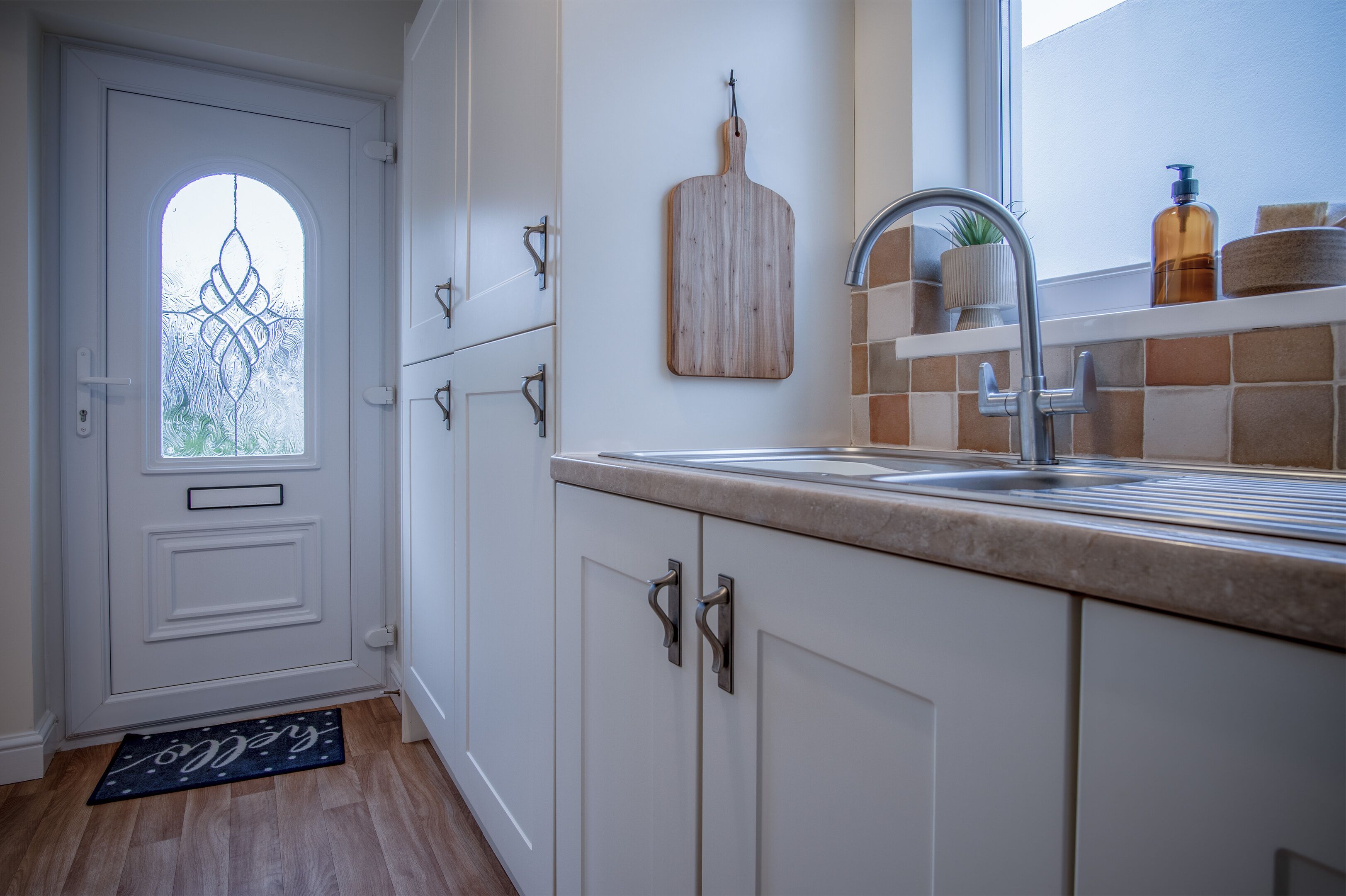 A kitchen with white cabinets, a sink, a wooden cutting board, and a window with frosted glass.