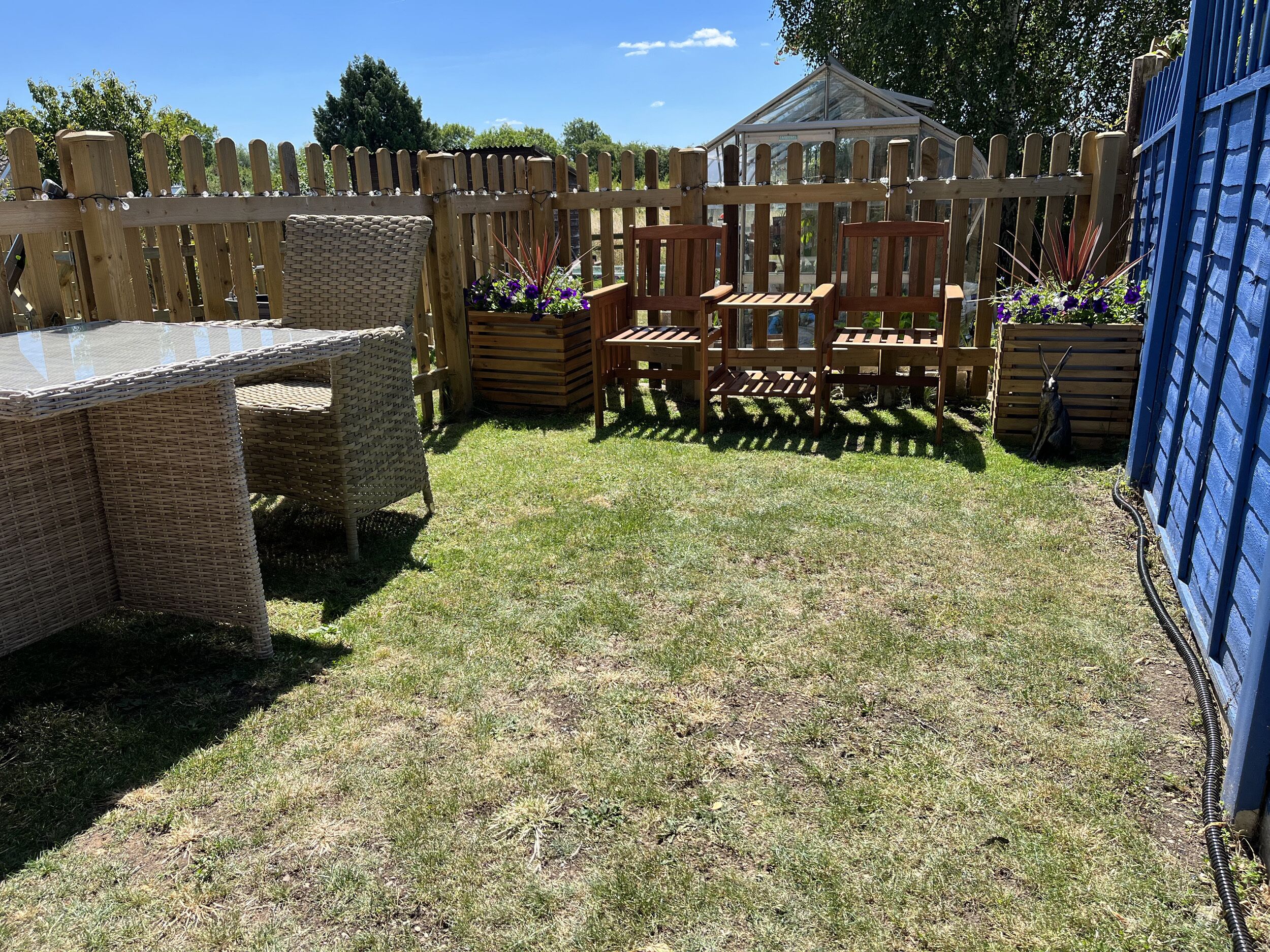 A backyard with a wooden fence, a glass greenhouse, wicker furniture, and a blue structure.