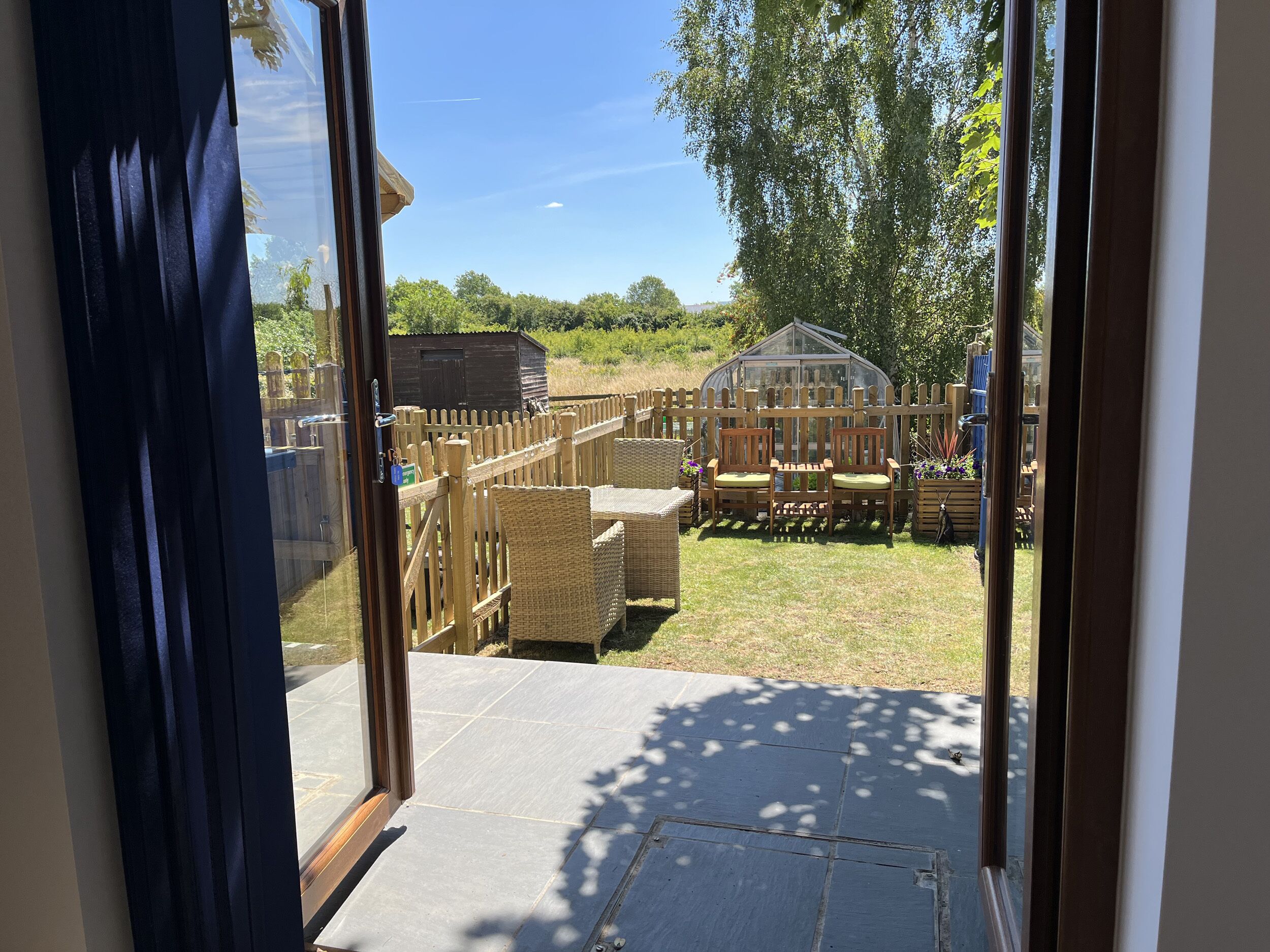 A view through a glass door into a backyard with a wooden fence, a greenhouse, and outdoor furniture.