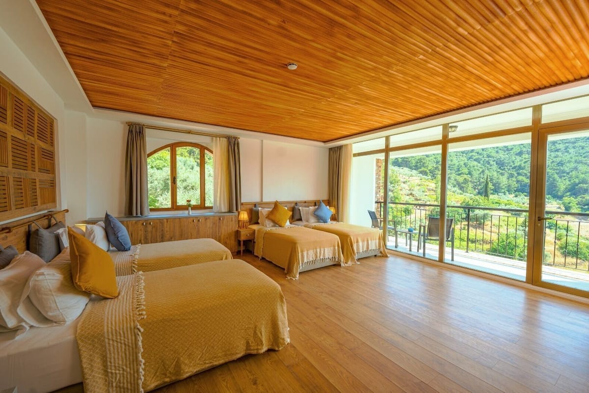 A room with two beds, wooden flooring, large windows with a view of greenery, and a wooden ceiling.