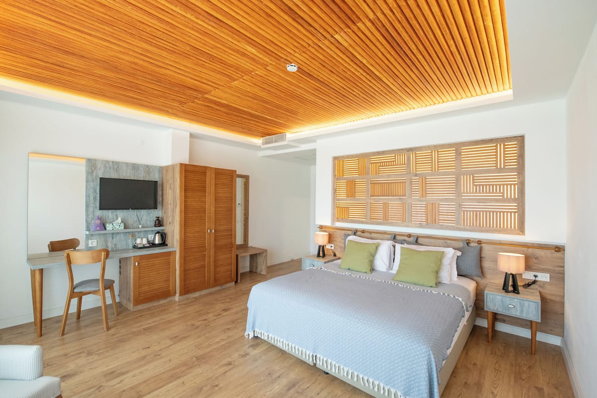 A modern bedroom with a wooden ceiling, a large bed with multiple pillows, a desk with a chair, and a TV mounted on the wall.