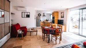 A modern living room with a dining area, a kitchen in the background, and a large sliding glass door to the outside. - Le Titibu: Gîte and Spa between Ardèche and Provence, ideal for two (Saint-Paulet-de-Caisson)