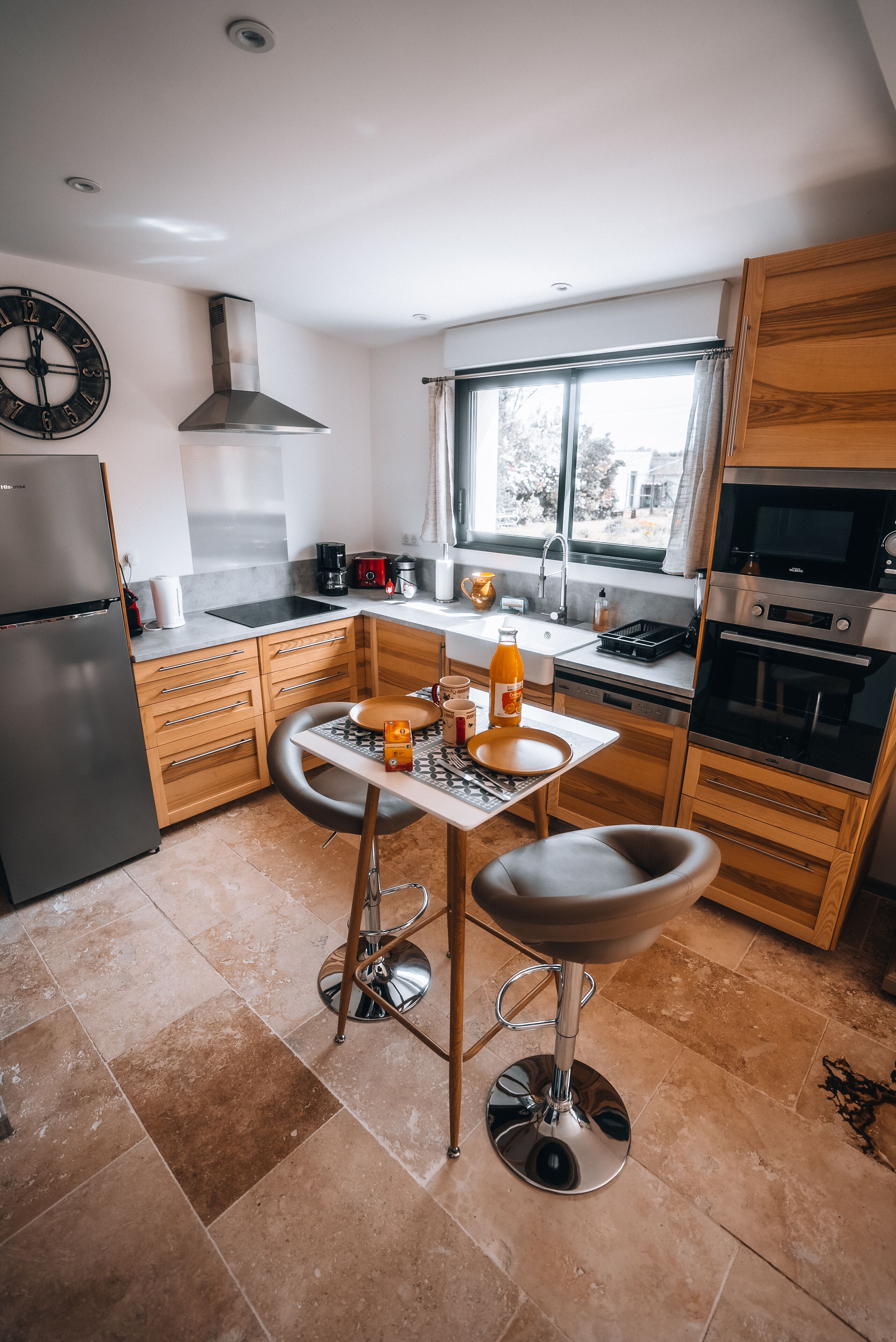 Een moderne keuken met een koelkast, fornuis, oven en houten keukenkastjes. Er is een raam met gordijnen, een spoelbak en een eethoek met krukken.