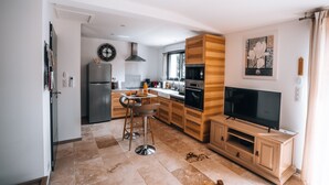 A modern kitchen with a dining area, wooden cabinets, a built-in oven, and a TV on a wooden stand. - Le Titibu: Gîte and Spa between Ardèche and Provence, ideal for two (Saint-Paulet-de-Caisson)