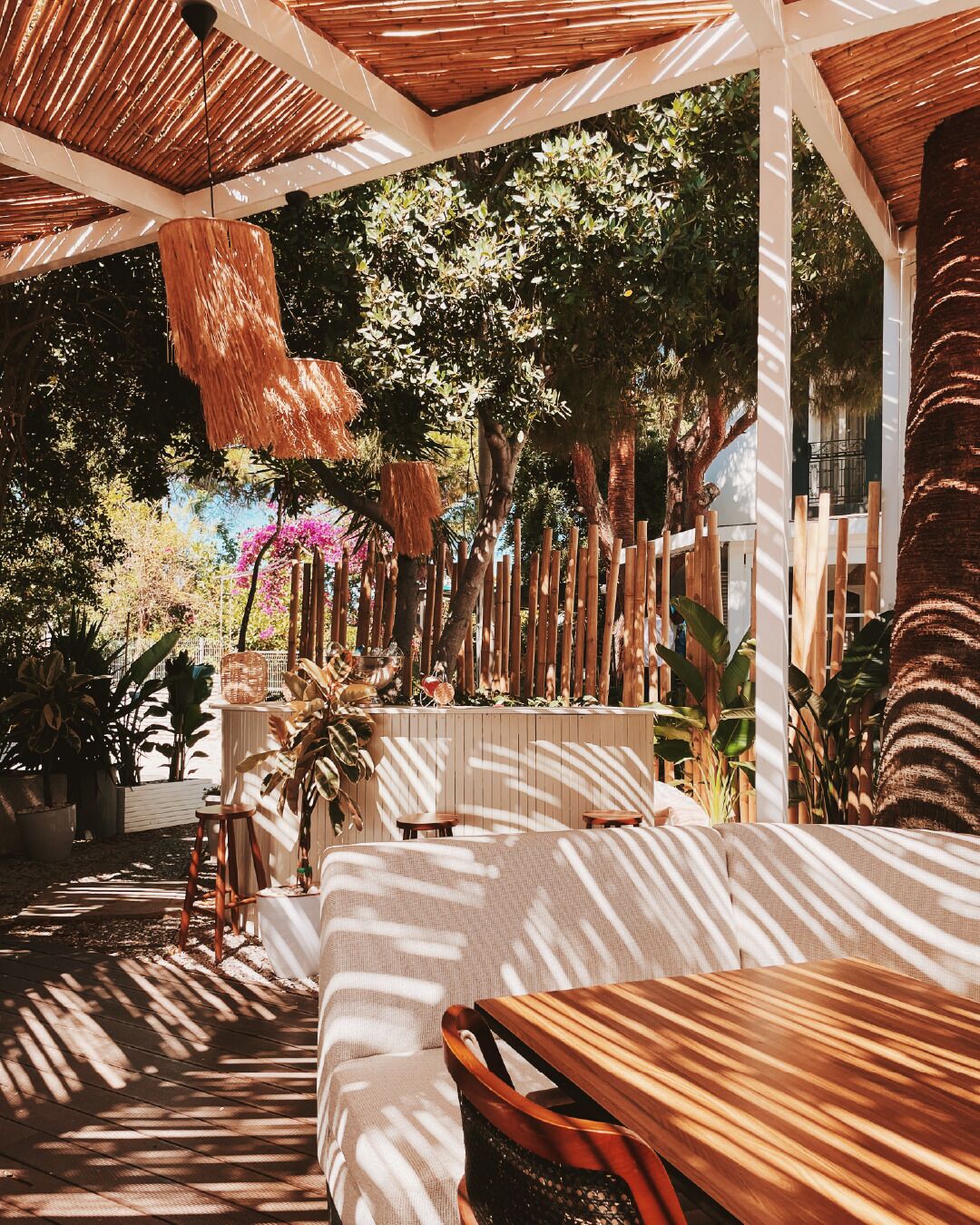 An outdoor seating area with wooden tables and chairs, surrounded by lush greenery and a bamboo fence.