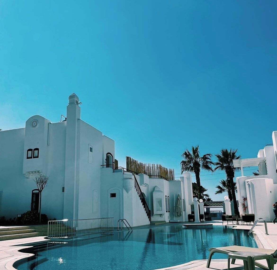A white building with a swimming pool, palm trees, and a clear blue sky.