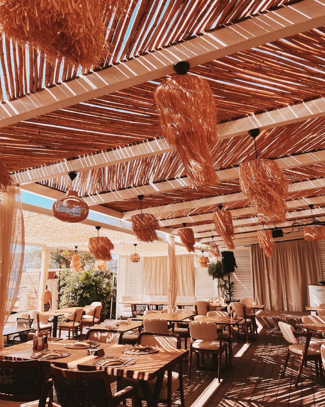 An outdoor dining area with bamboo ceiling structures, hanging lights, and a variety of seating arrangements.