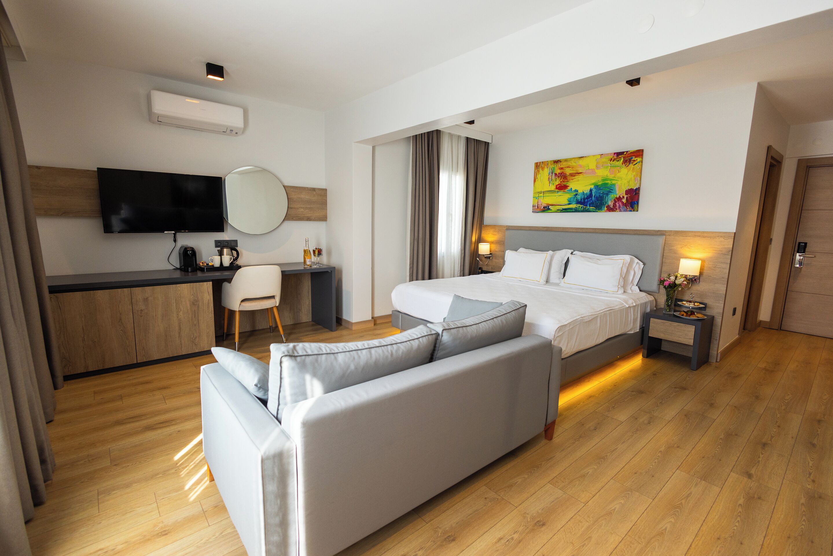 A modern hotel room with a bed, a sofa, a desk, a television, and a painting on the wall.