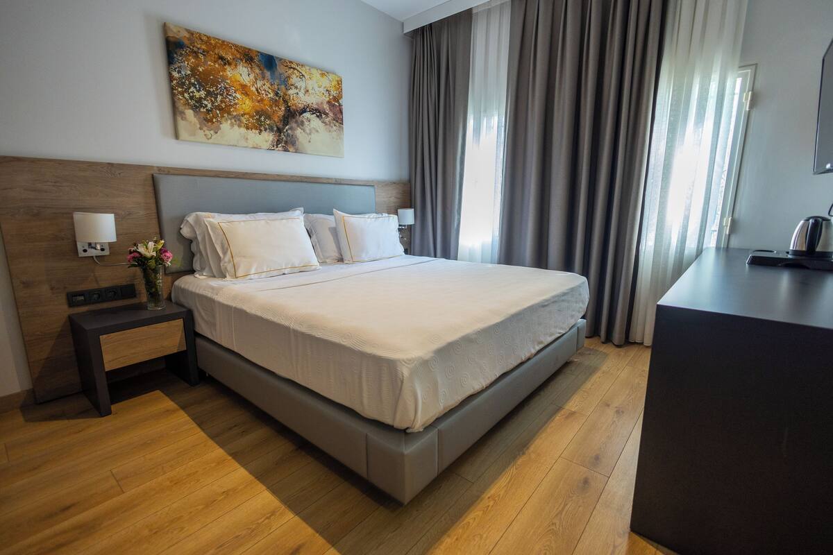 A modern hotel room with a large bed, a bedside table with a lamp, a desk, and a painting on the wall.