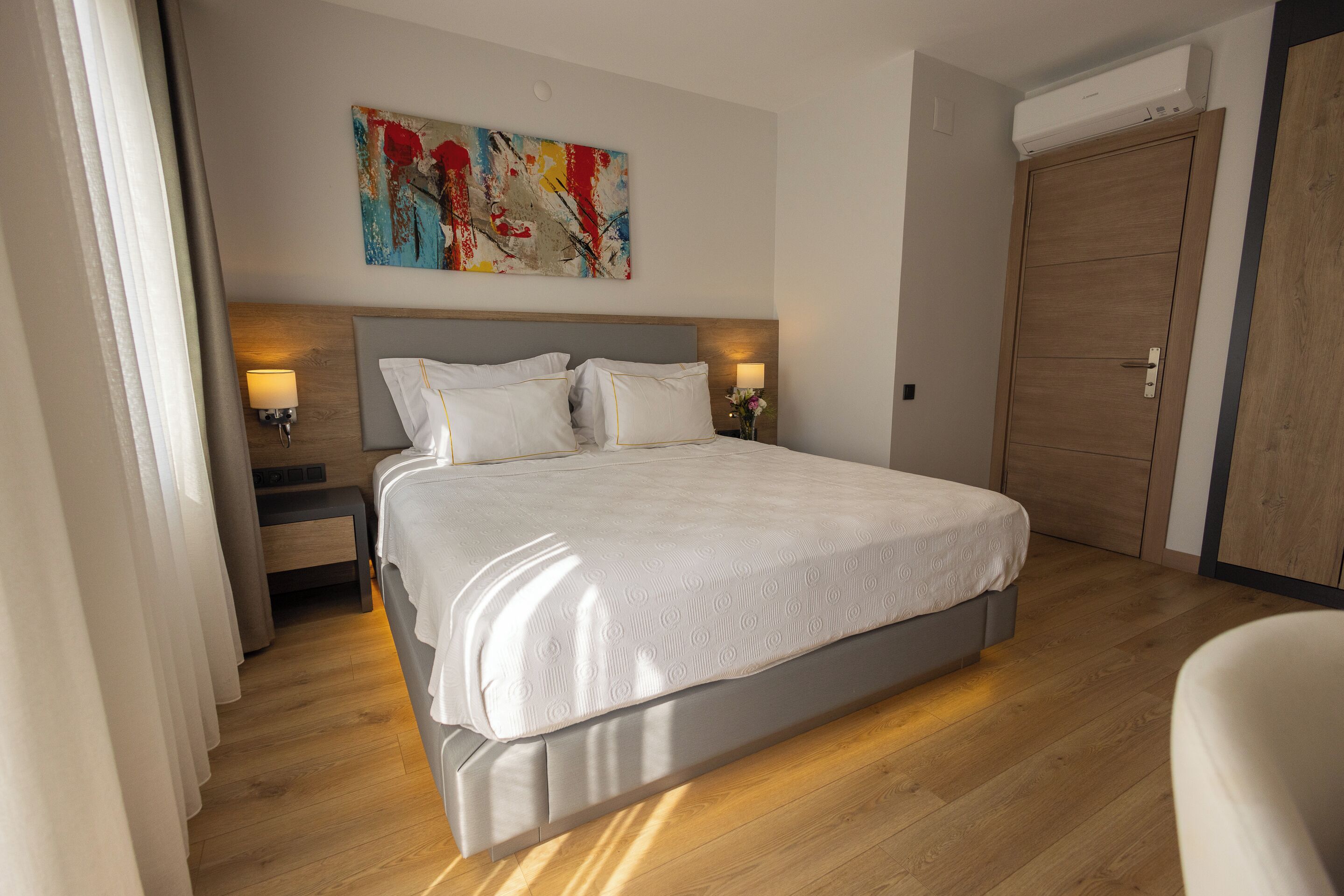 A hotel room with a large bed, two bedside lamps, a wooden headboard, a colorful abstract painting, and a wooden wardrobe.