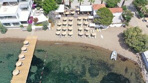 Eine Vogelperspektive zeigt ein Hotel am Strand mit einer Holzterrasse, Liegestühlen und Sonnenschirmen.