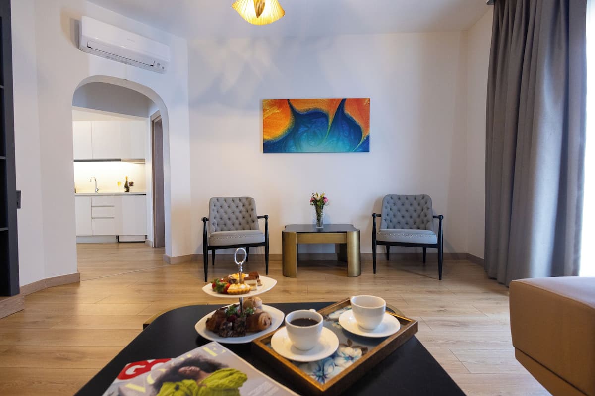 A modern hotel room with a dining area, two armchairs, a small table with a vase, and a tray with food and drinks.