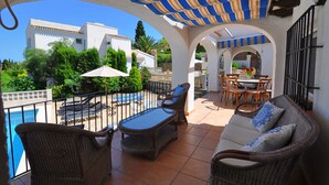 Un patio cubierto al aire libre con mobiliario de mimbre, piscina y comedor.