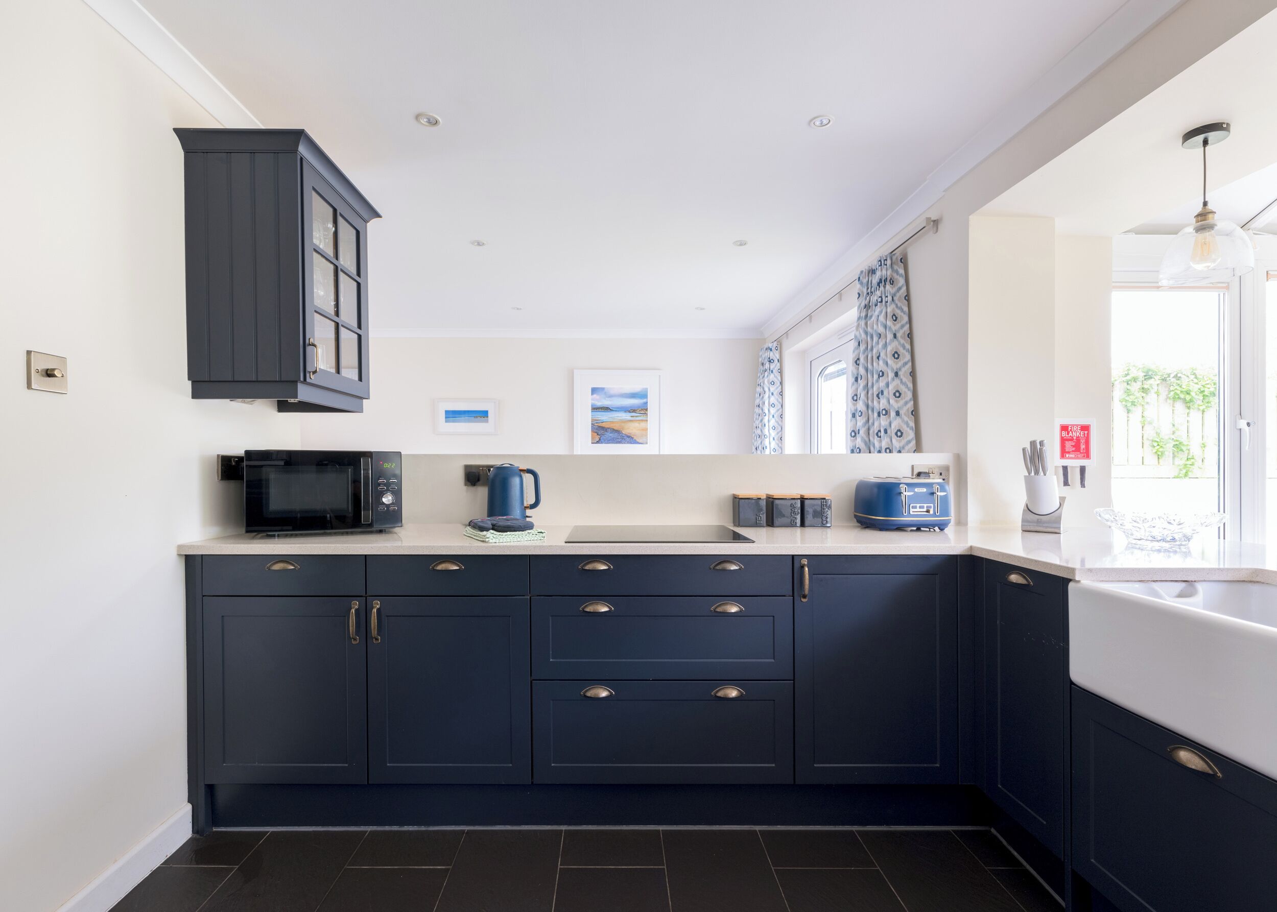 A modern kitchen with dark blue cabinets, a microwave, a kettle, and a toaster. There are framed pictures on the wall and a window with curtains.