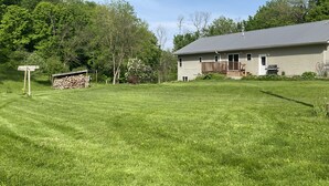 A single-story house with a covered porch, a stack of firewood, and a wooden signpost in a grassy yard. - Private vacation house for larger groups near the Dells with heated game room. (Reedsburg)
