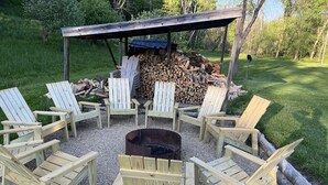 A fire pit surrounded by six Adirondack chairs, with a pile of firewood and a wooden shelter in the background. - Private vacation house for larger groups near the Dells with heated game room. (Reedsburg)