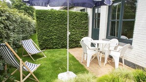 Een terras met een tafel en stoelen, een gestreept lounge stoel en een blauwe parasol.