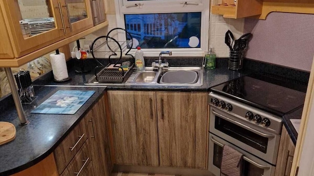 A compact kitchen with wooden cabinets, a black countertop, a stove, and a window with a view of a building. - Discover Comfort Home From Home 8-birth Caravan (Skegness)