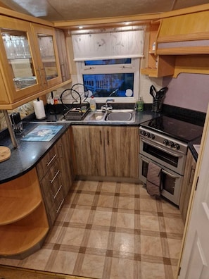 A compact kitchen with wooden cabinets, a black countertop, a stove, and a window with a view of a building. - Discover Comfort Home From Home 8-birth Caravan (Skegness)