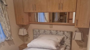 A bedroom with a bed, wooden cabinets, a mirror, and two bedside lamps. - Discover Comfort Home From Home 8-birth Caravan (Skegness)