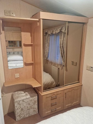 A compact bedroom with a wooden wardrobe, a small bedside table, a single bed, and a mirror. - Discover Comfort Home From Home 8-birth Caravan (Skegness)